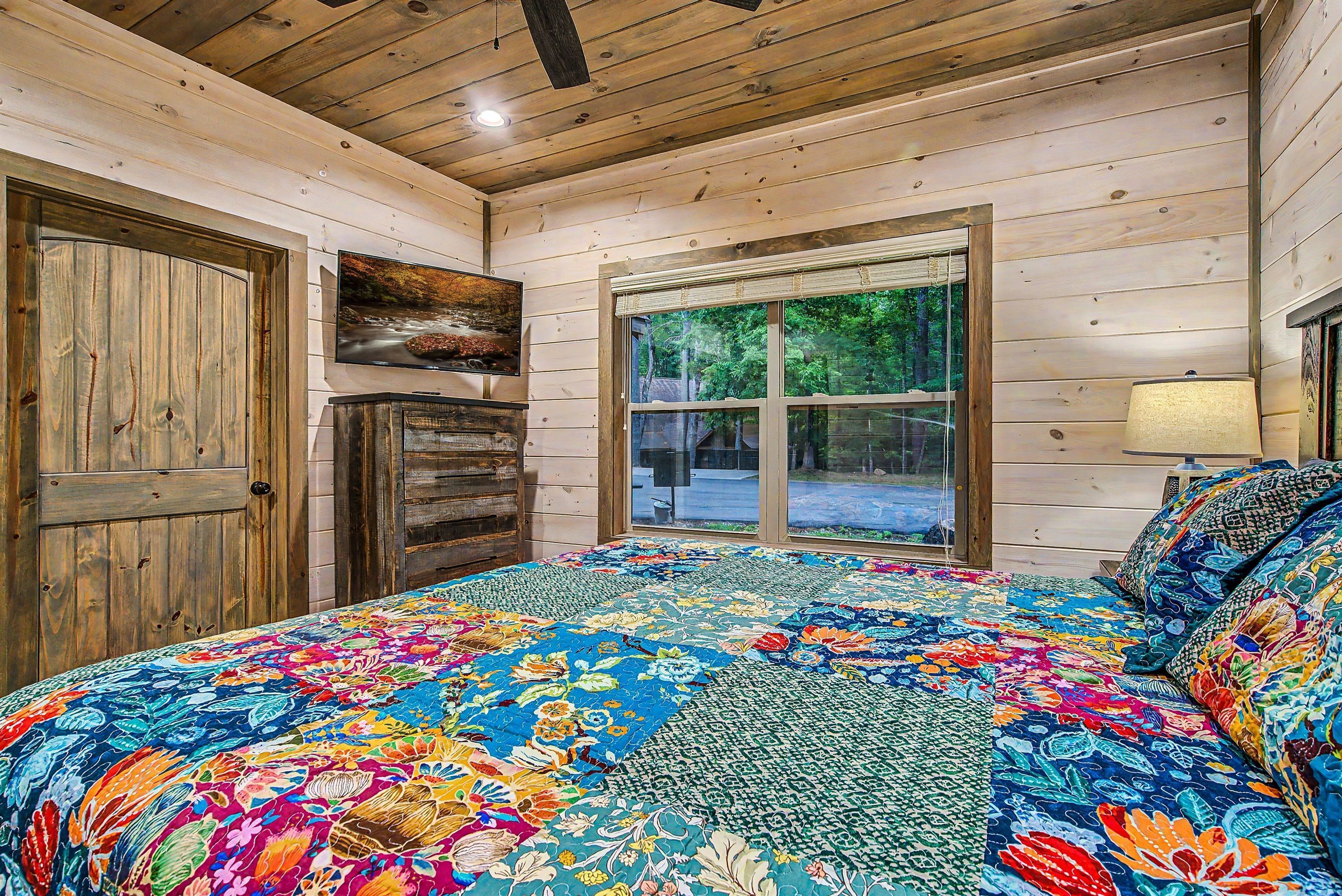 A room with a wooden ceiling, a large bed with a colorful quilt, a wooden door, and a window with a view of trees.
