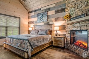 Un dormitorio con una cama, una mesita de noche, una chimenea de piedra y una pared con paneles de madera y un cuadro.