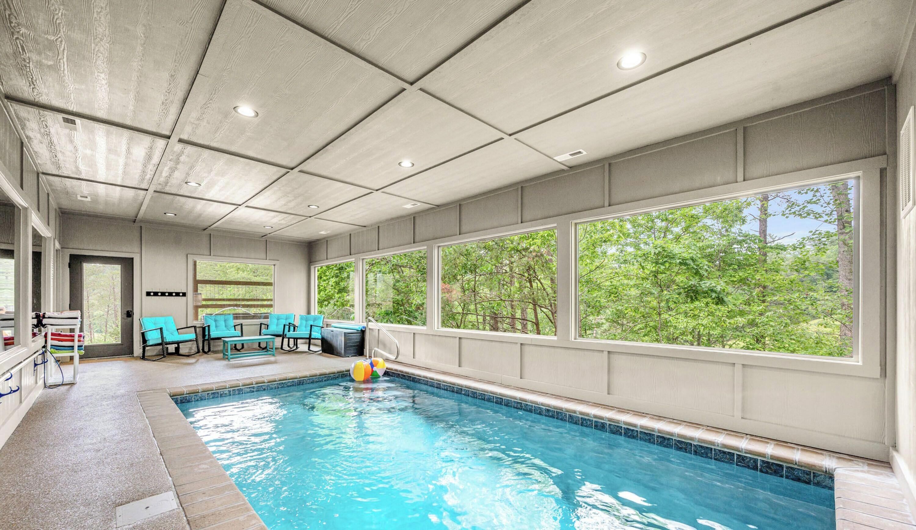 An indoor swimming pool with clear windows, a seating area with chairs, and a view of greenery outside.