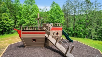 A playground with a ship-themed play structure, including a slide and climbing steps, surrounded by trees.