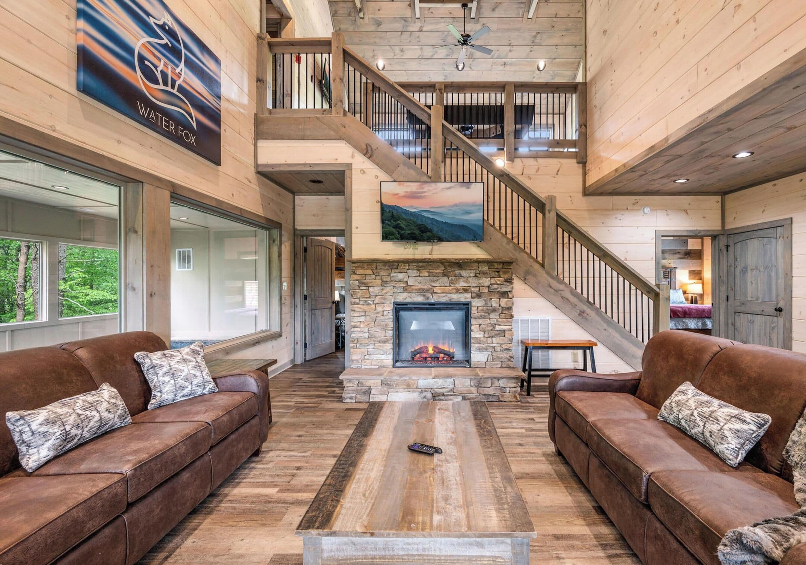 A spacious interior with a stone fireplace, leather sofas, a wooden staircase, and a large window overlooking greenery.