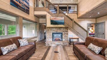 A spacious interior with a stone fireplace, leather sofas, a wooden staircase, and a large window overlooking greenery.