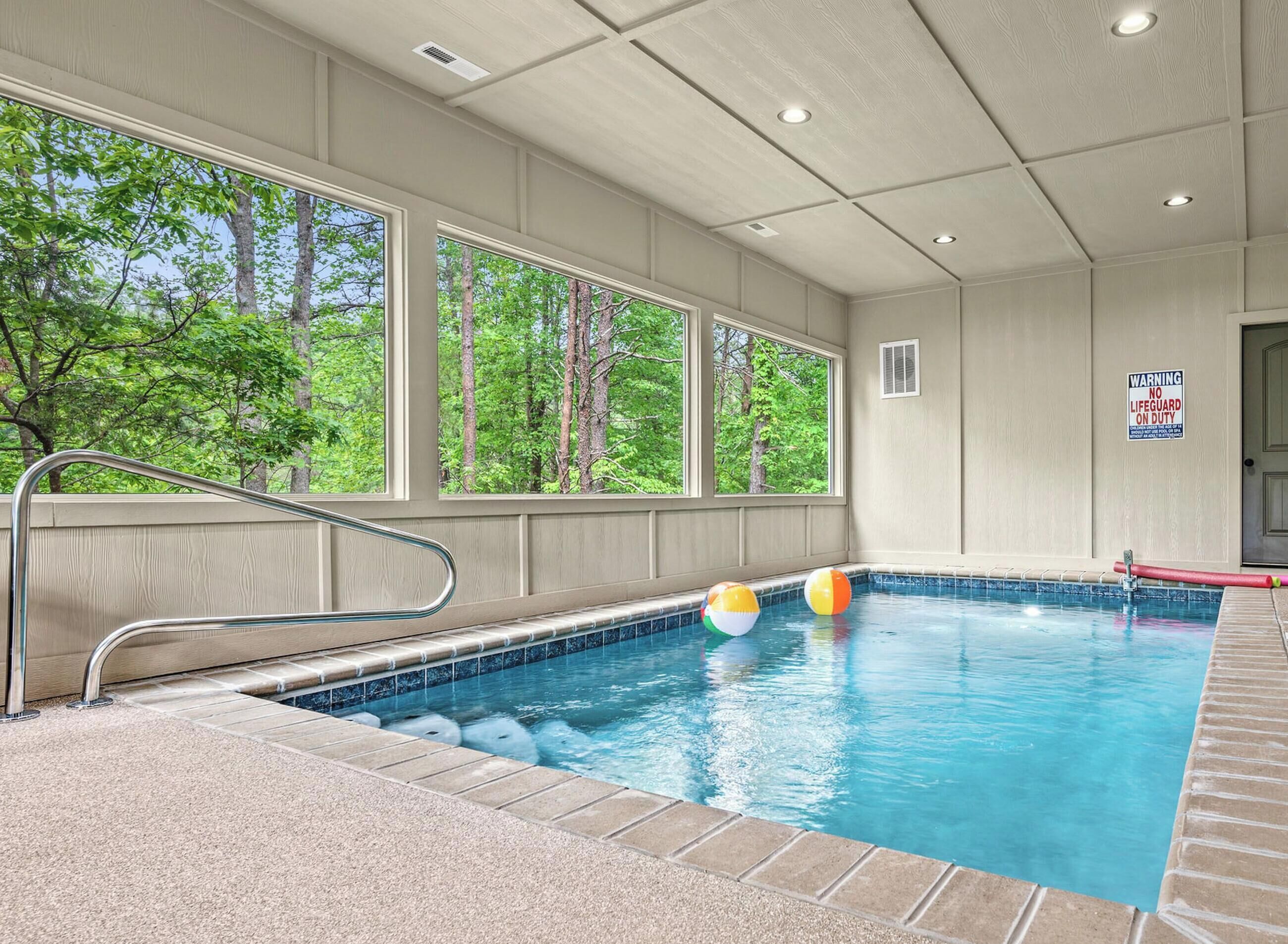 An indoor swimming pool with a view of trees through large windows, a lifeguard sign, and a poolside area with a red barrier.