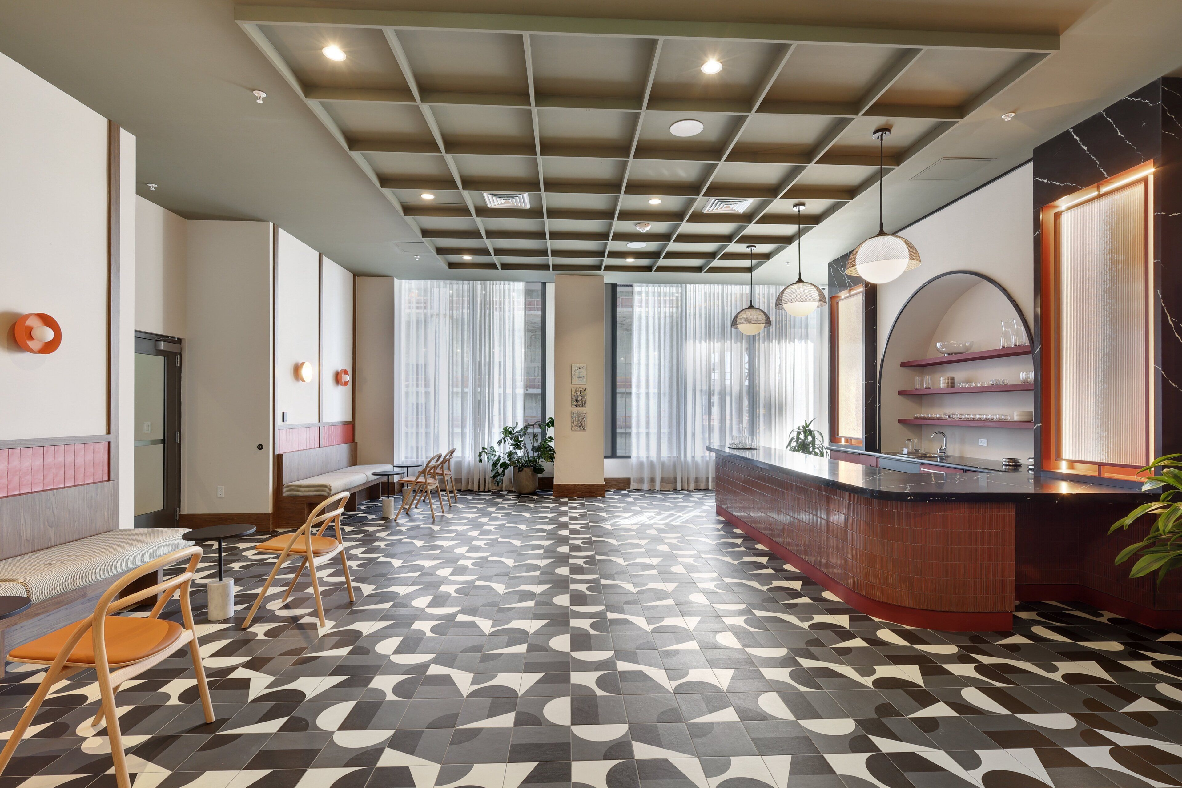 Un hall d’hôtel moderne avec un sol à motifs géométriques, un comptoir d’accueil, un espace salon et de grandes fenêtres ornées de rideaux légers.