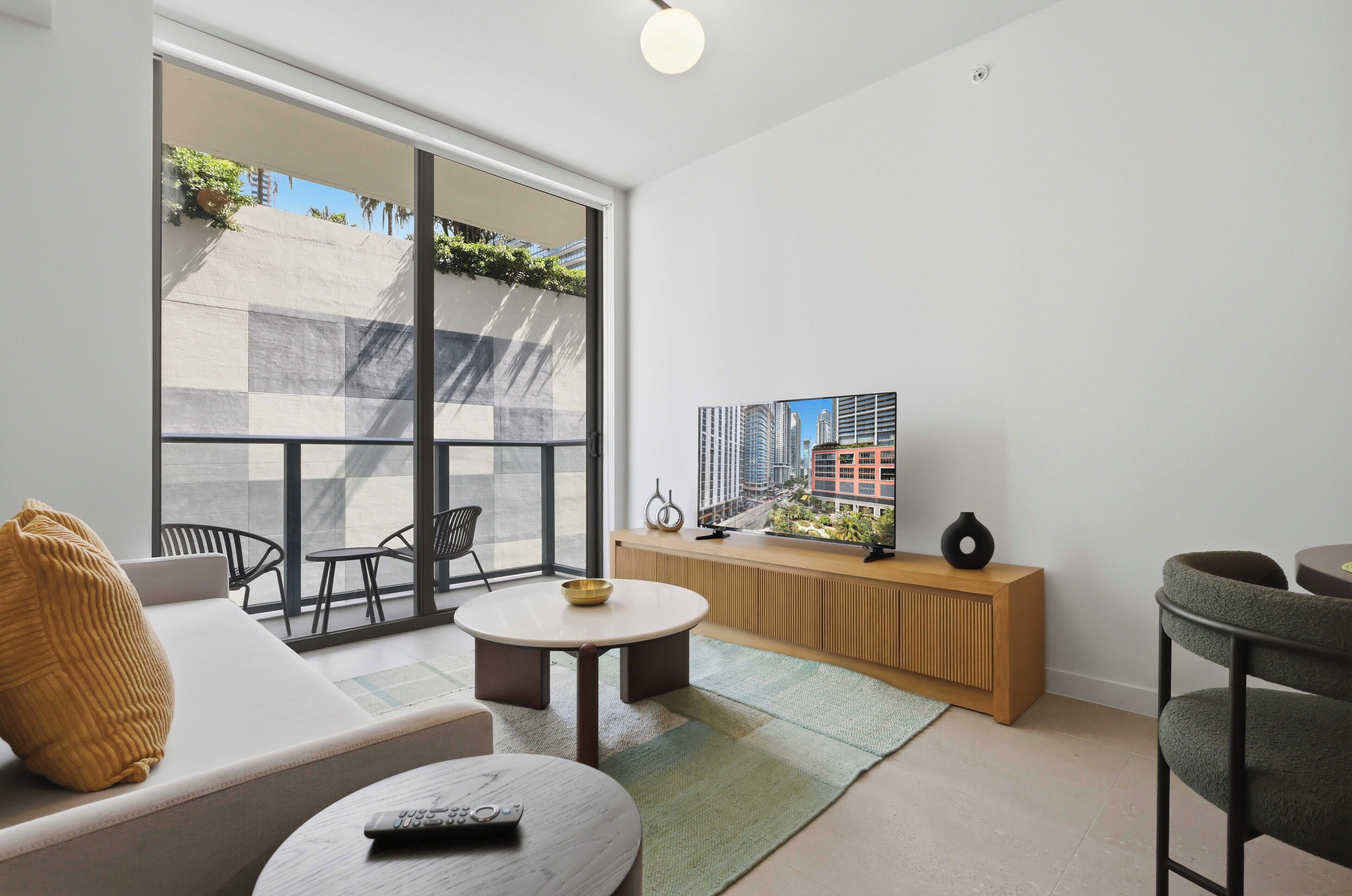 Una sala moderna con un sofá, una mesa de centro, un televisor y vistas a un paisaje urbano a través de amplios ventanales.