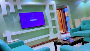 A room with a flat-screen TV mounted on a white wall, teal armchairs, and a marble coffee table.
