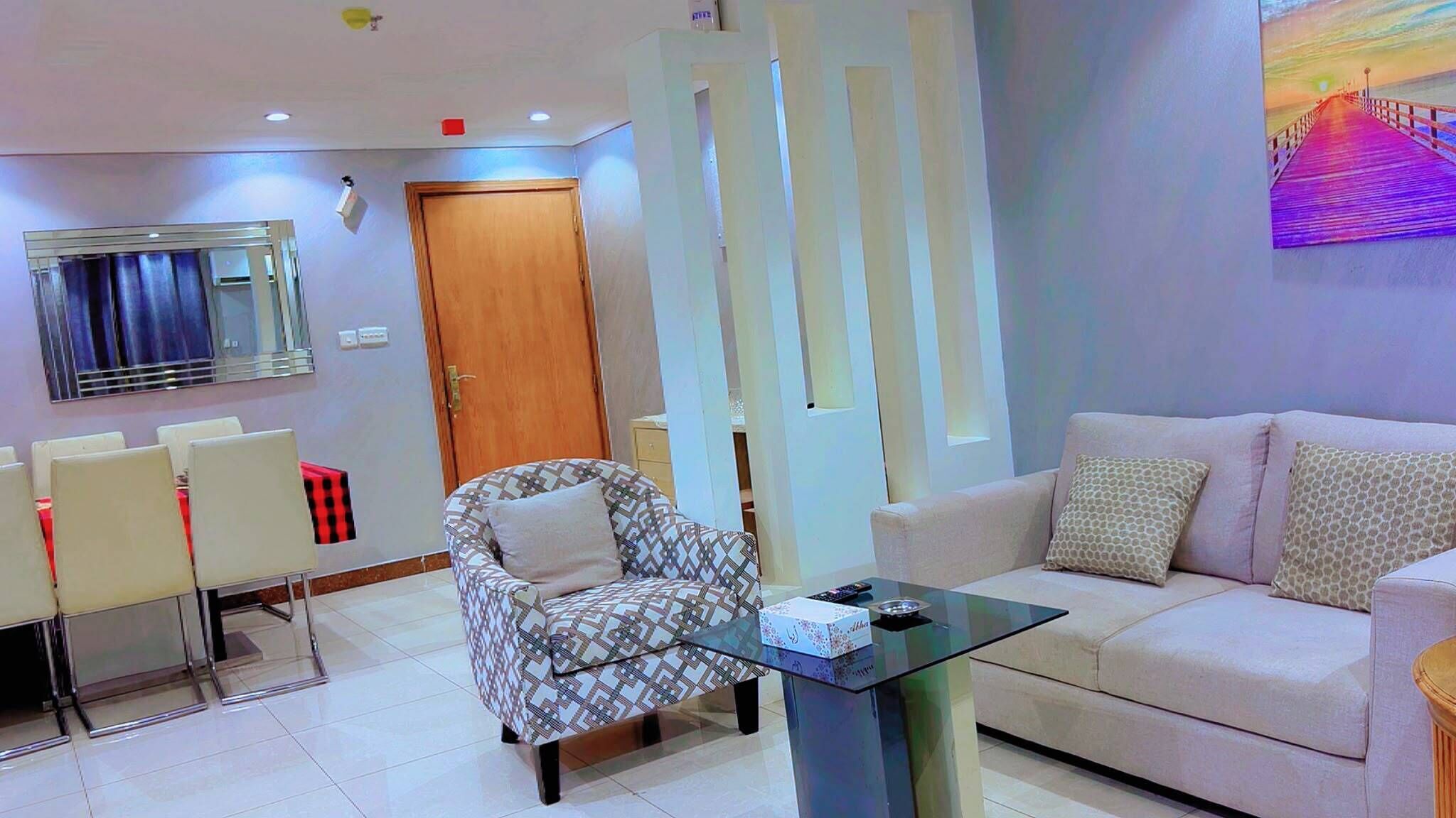 A modern living room with a sofa, armchairs, a glass coffee table, and a dining area with chairs.
