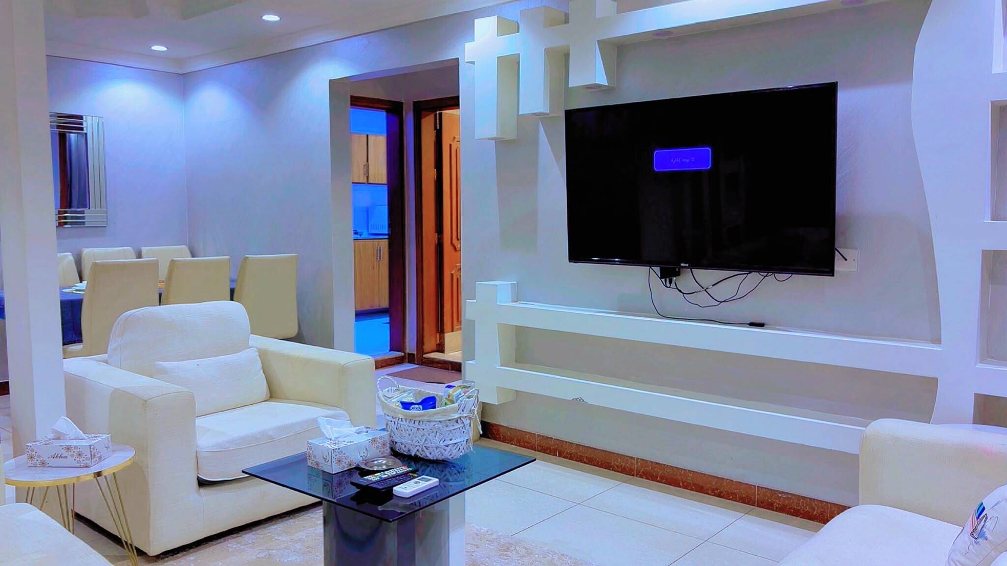 Een moderne woonkamer met een flatscreen televisie aan de muur, een bank, een salontafel en een eethoek op de achtergrond.