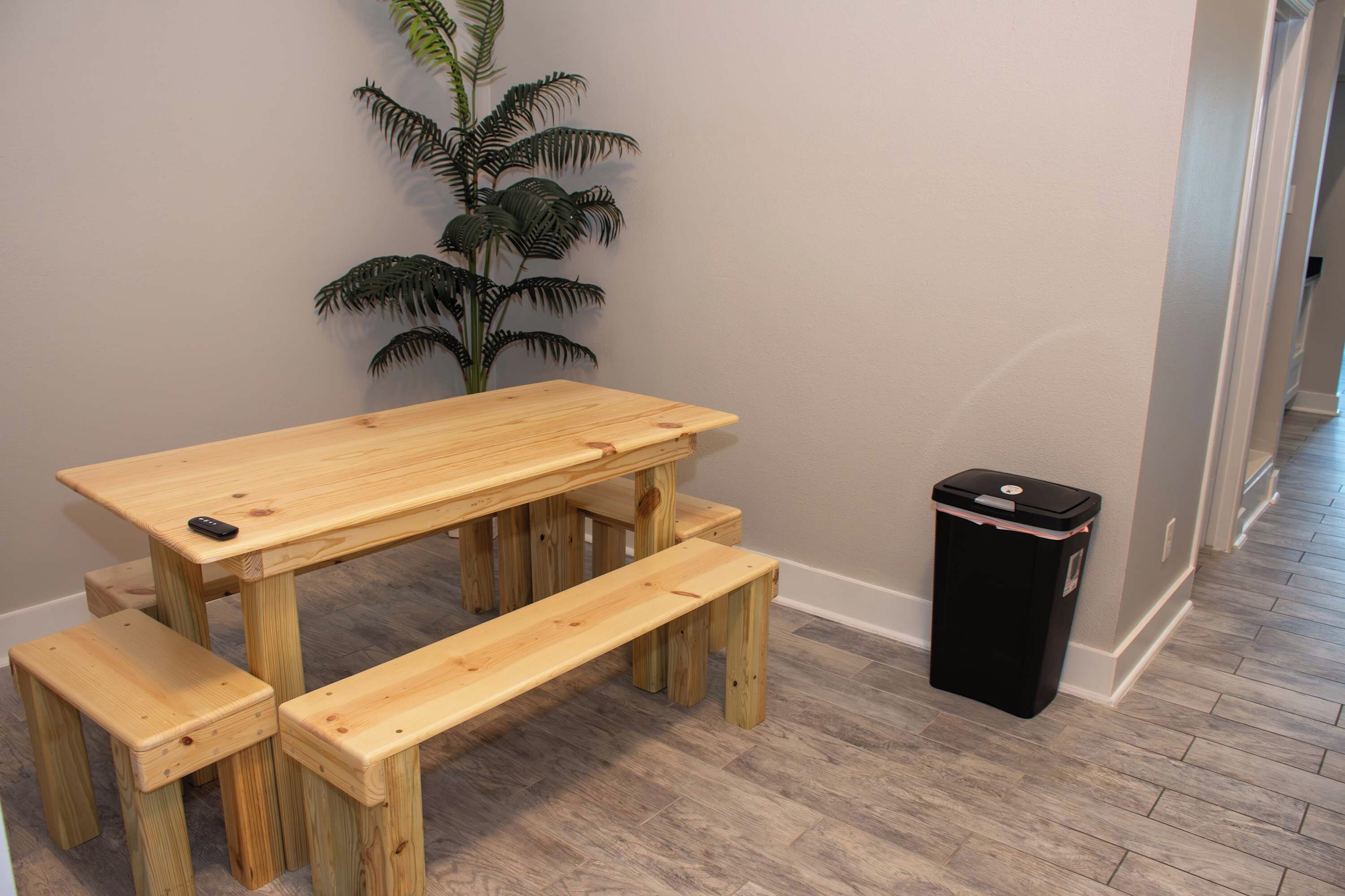A wooden table with benches in a room with a plant and a trash bin.