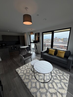 Ein modernes Wohnzimmer mit einer Couch, zwei runden Tischen, einem großen Fenster mit Blick auf die Stadt und einer Küchenzeile.