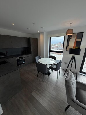 A modern kitchen with dark cabinetry, a round dining table with chairs, and a large window offering a city view. - Stunning 2 Bedroom Apartment-2 Bath with Balcony (Salford)
