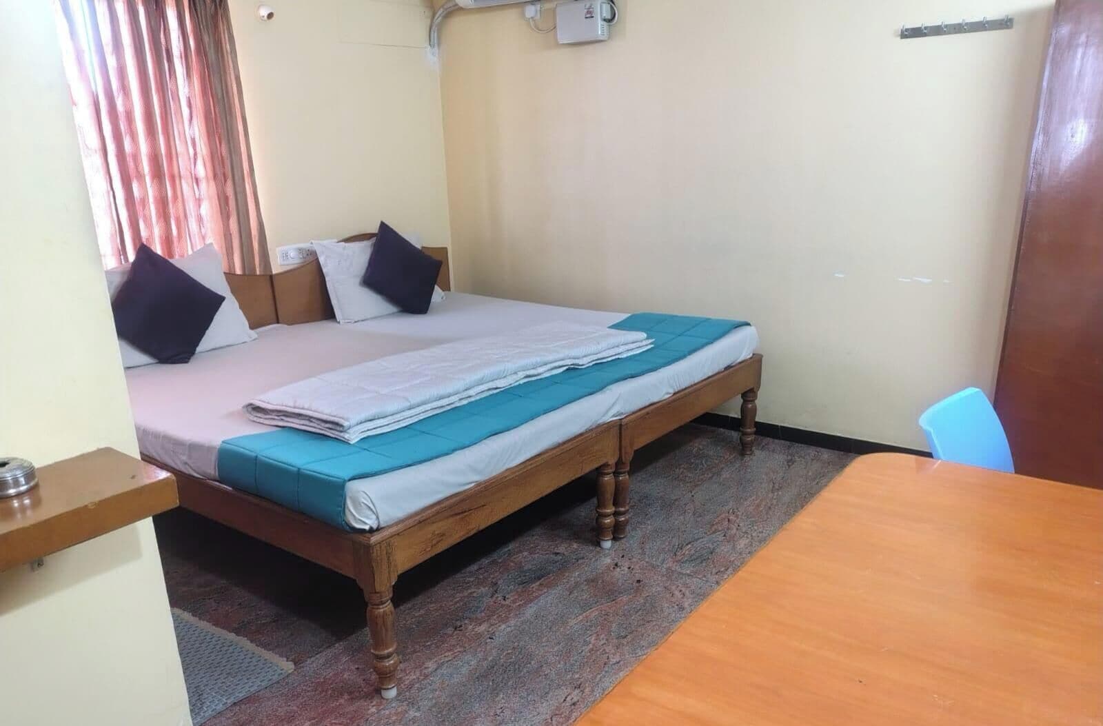 A single bed with a wooden frame, a blue mattress cover, and a white blanket. There is a wooden table with a chair, a small shelf, and a wall-mounted electrical outlet.