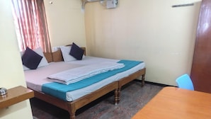 A single bed with a wooden frame, a blue mattress cover, and a white blanket. There is a wooden table with a chair, a small shelf, and a wall-mounted electrical outlet. - Hotel Royal Park (Coimbatore)