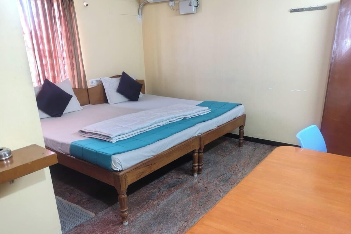 A single bed with a wooden frame, a blue mattress cover, and a white blanket. There is a wooden table with a chair, a small shelf, and a wall-mounted electrical outlet.