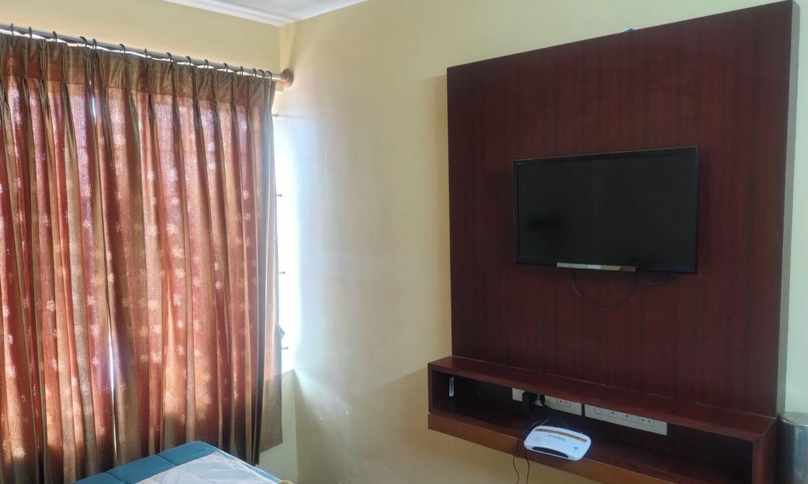 Una habitación de hotel con televisión instalada en un panel de madera, una ventana con cortinas y una mesita de noche.
