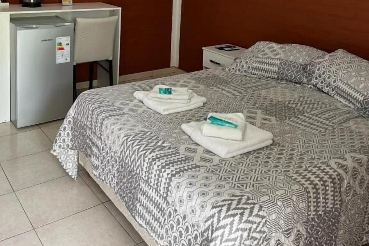 A hotel room with a bed, a desk with a kettle, and an air conditioning unit.