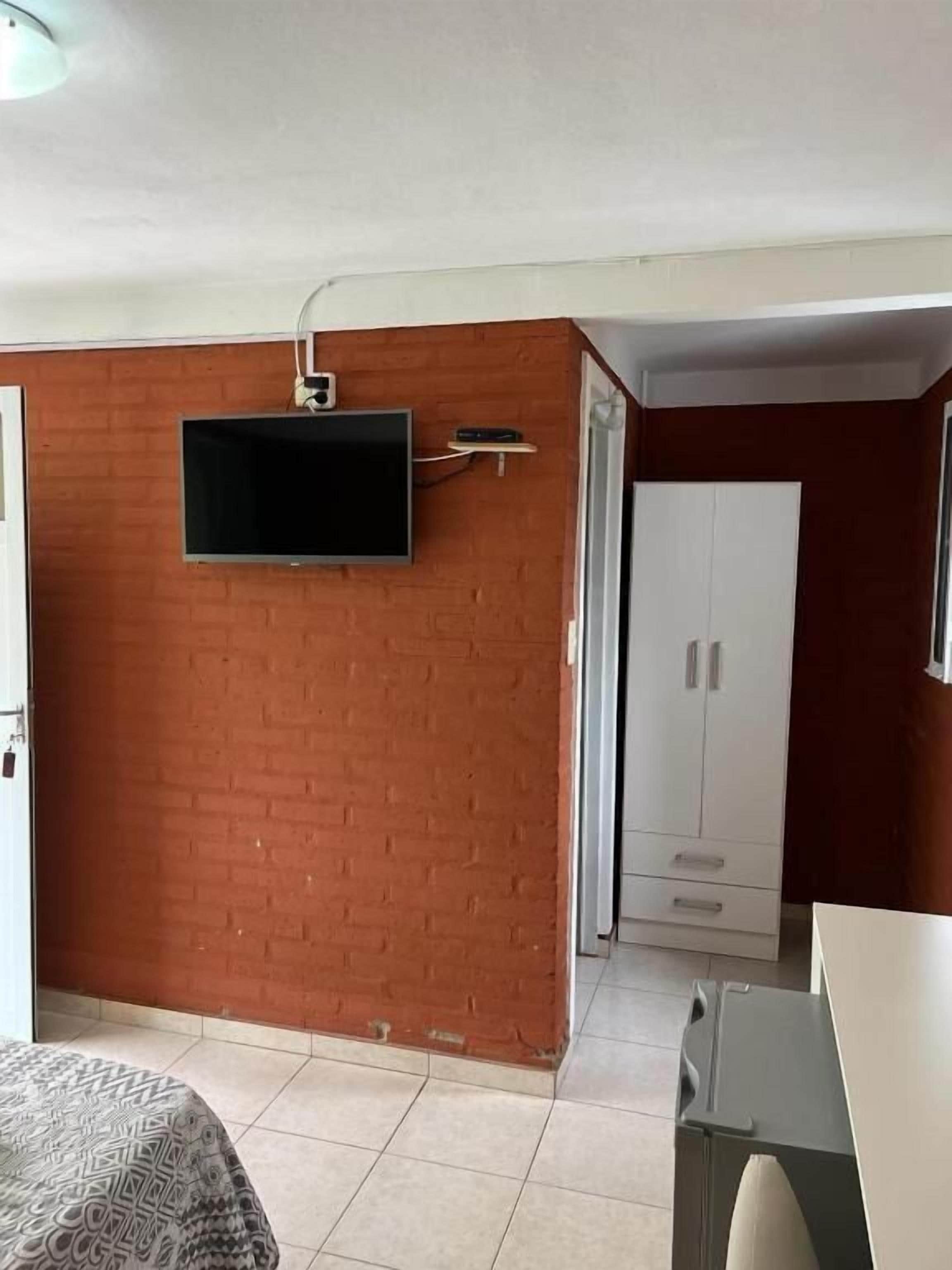 Une pièce avec un téléviseur fixé au mur en briques, une armoire et un lit recouvert d’une couvre-lit à motifs.