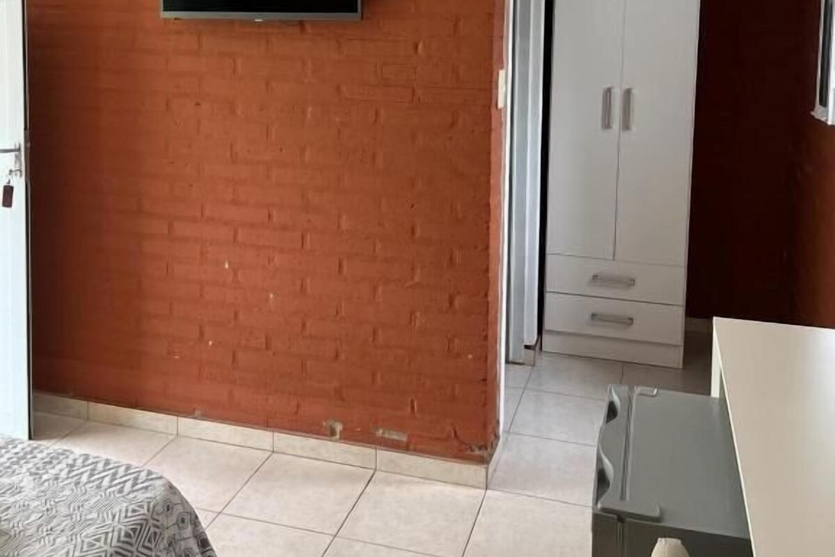 A room with a television mounted on a brick wall, a wardrobe, and a bed with a patterned bedspread.