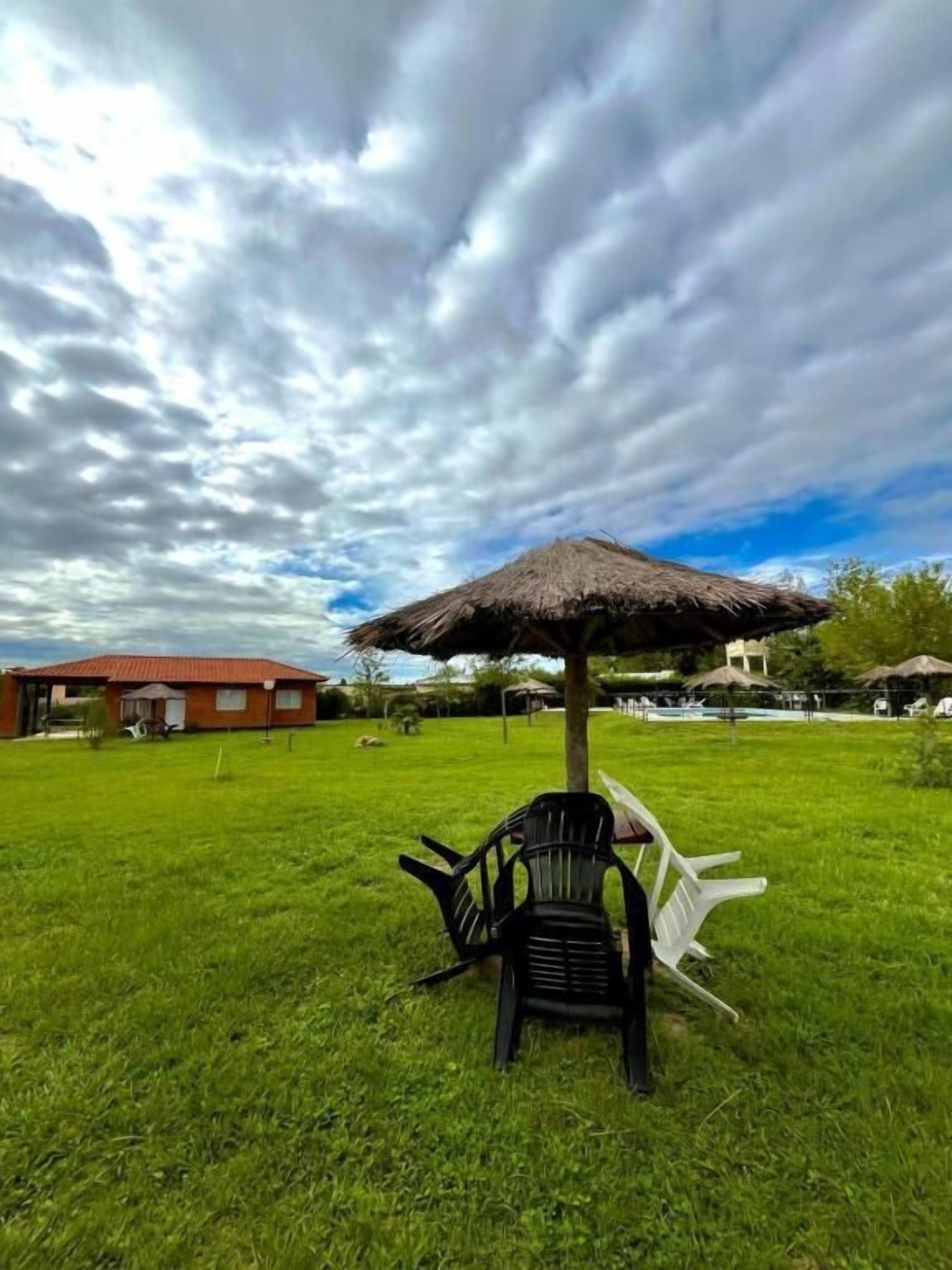 A grassy area with a thatched umbrella, two black chairs, and a red building in the background.