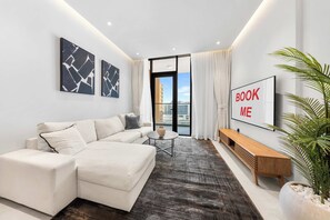 A modern living room with a sectional sofa, a coffee table, a TV, and a large window with a city view. - StayShort - Bright & Roomy 2 bed with Balcony that Sleeps 6 (Majan)