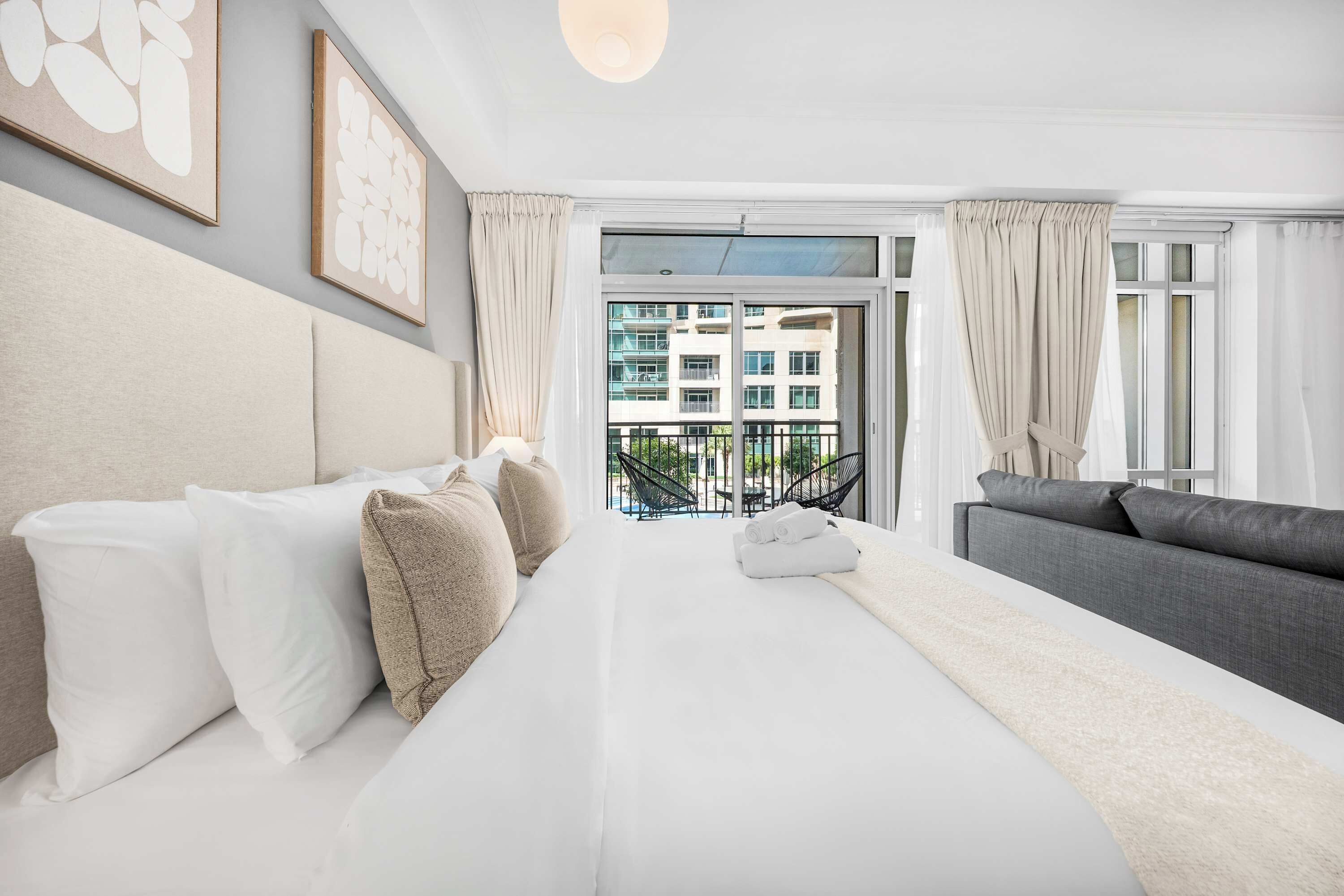 A hotel room with a bed, a sofa, a balcony with a view of buildings, and a ceiling light.