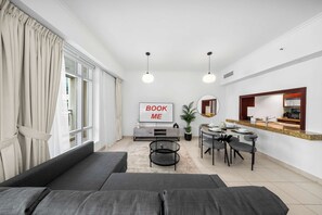 Ein modernes Wohnzimmer mit einer Couch, einem Essbereich mit Tisch für vier Personen, einem Fernseher mit der Aufschrift 'BOOK ME' und einer durch eine Glastrennwand sichtbaren Küche.