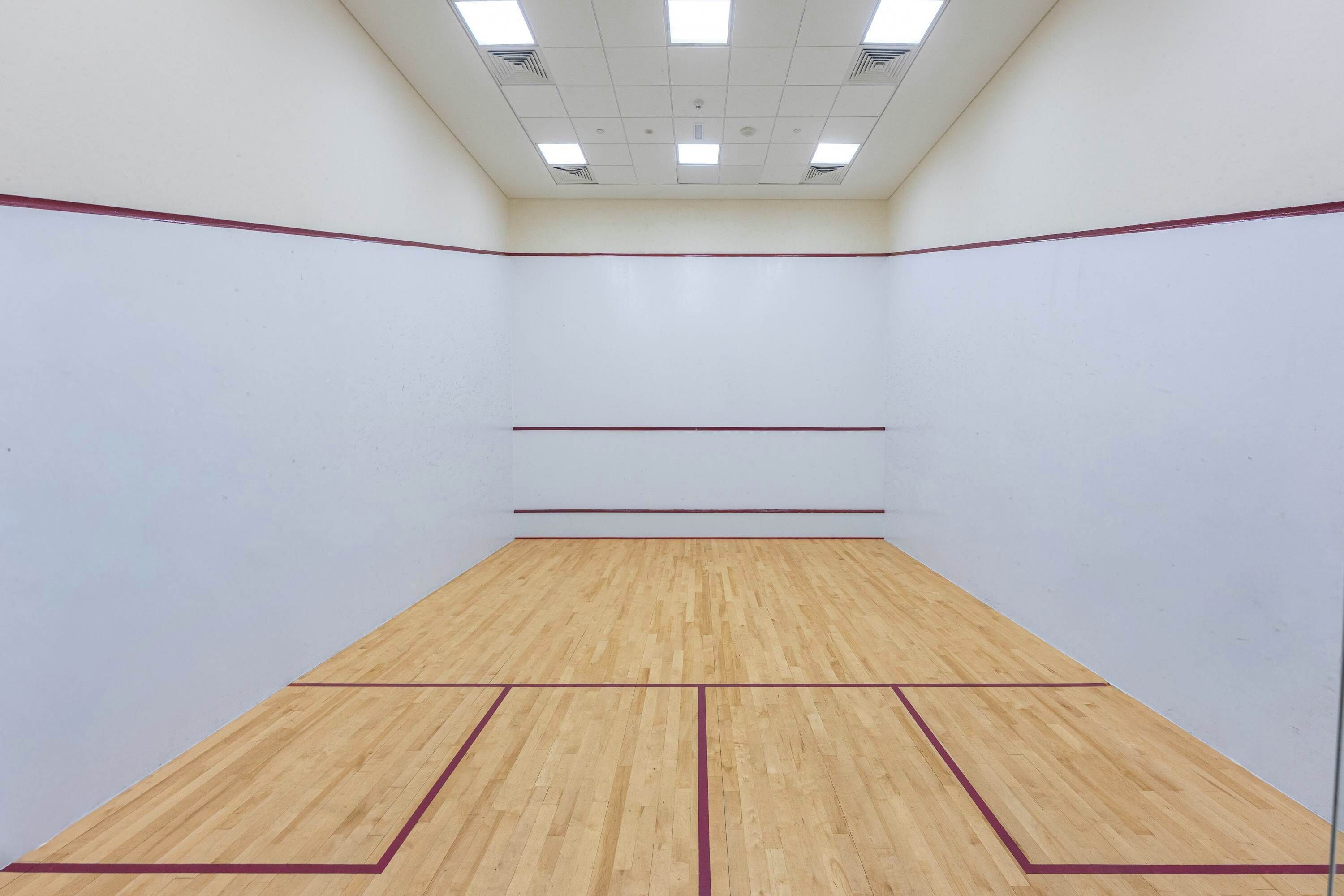 A squash court with a wooden floor and white walls, equipped with ceiling lights.