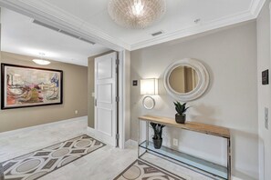 A hallway with a patterned floor, a framed abstract painting, a door, a wall-mounted mirror, a console table with a plant, and a light fixture. - Grande Geneva at The Dunes (Naples)