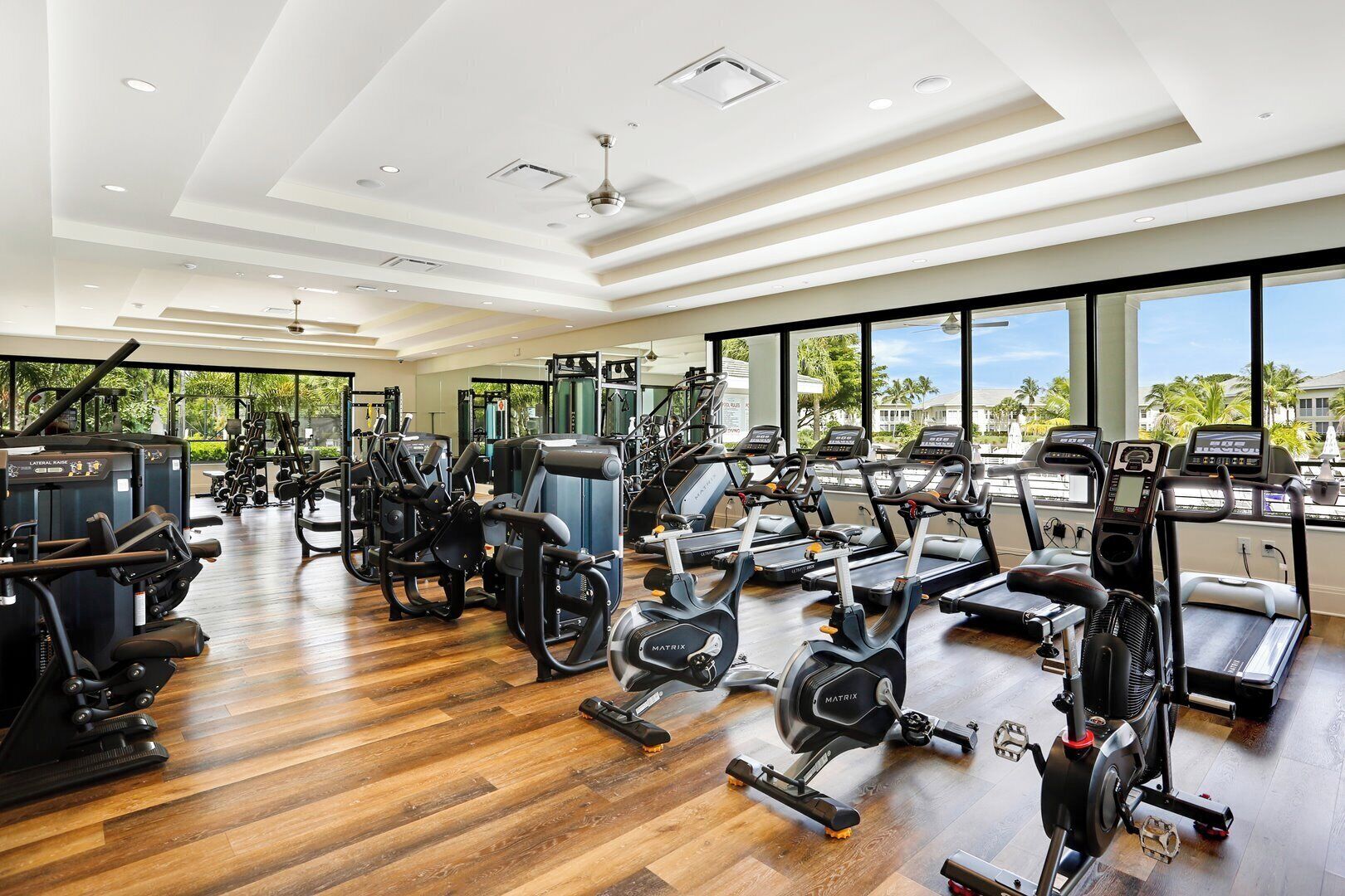 A spacious gym with various exercise equipment, large windows, and a wooden floor.
