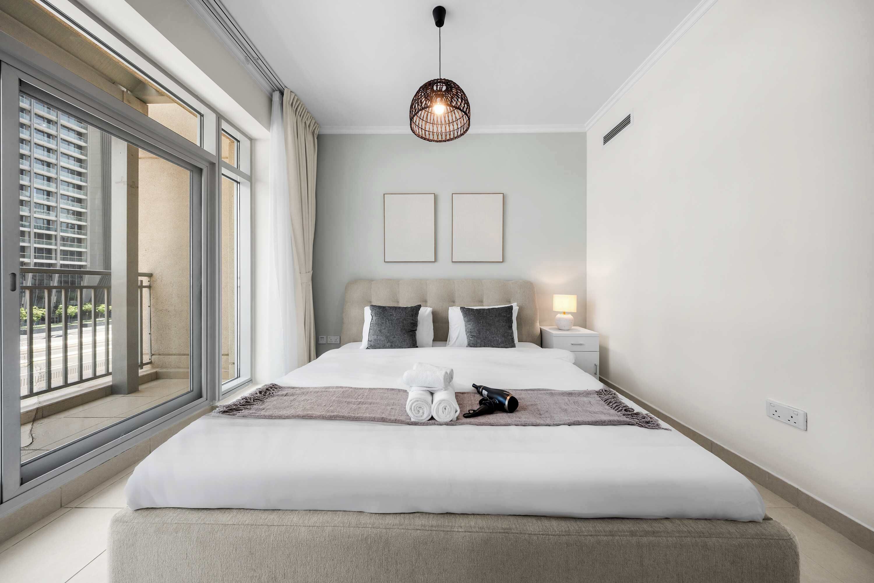 Habitación de hotel moderna con una cama grande, vistas de la ciudad a través de ventanales de piso a techo, una lámpara colgante y una mesita de noche con lámpara.