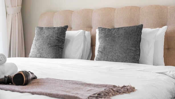 Ein ordentlich bezogenes Bett mit weißen und grauen Kissen, einer braunen Tagesdecke und einem zusammengerollten Handtuch.