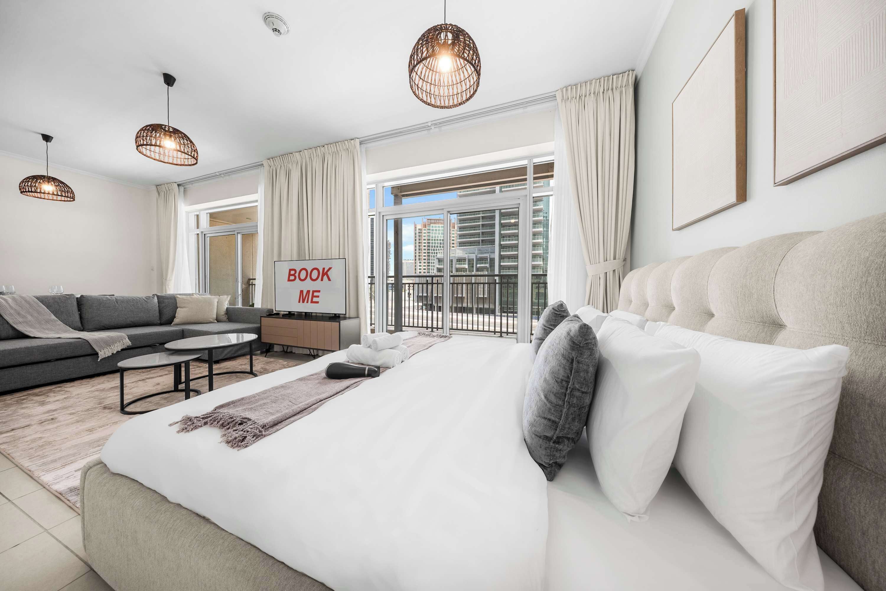A modern hotel room with a large bed, a sofa, a coffee table, and a TV. There is a sign that says 'BOOK ME' on the wall.
