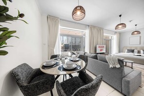 A modern living room with a dining area, grey sofas, a dining table set with plates and glasses, and a 'BOOK ME' sign. - StayShort - Luxury Studio Apartment in Downtown with Balcony (Downtown)