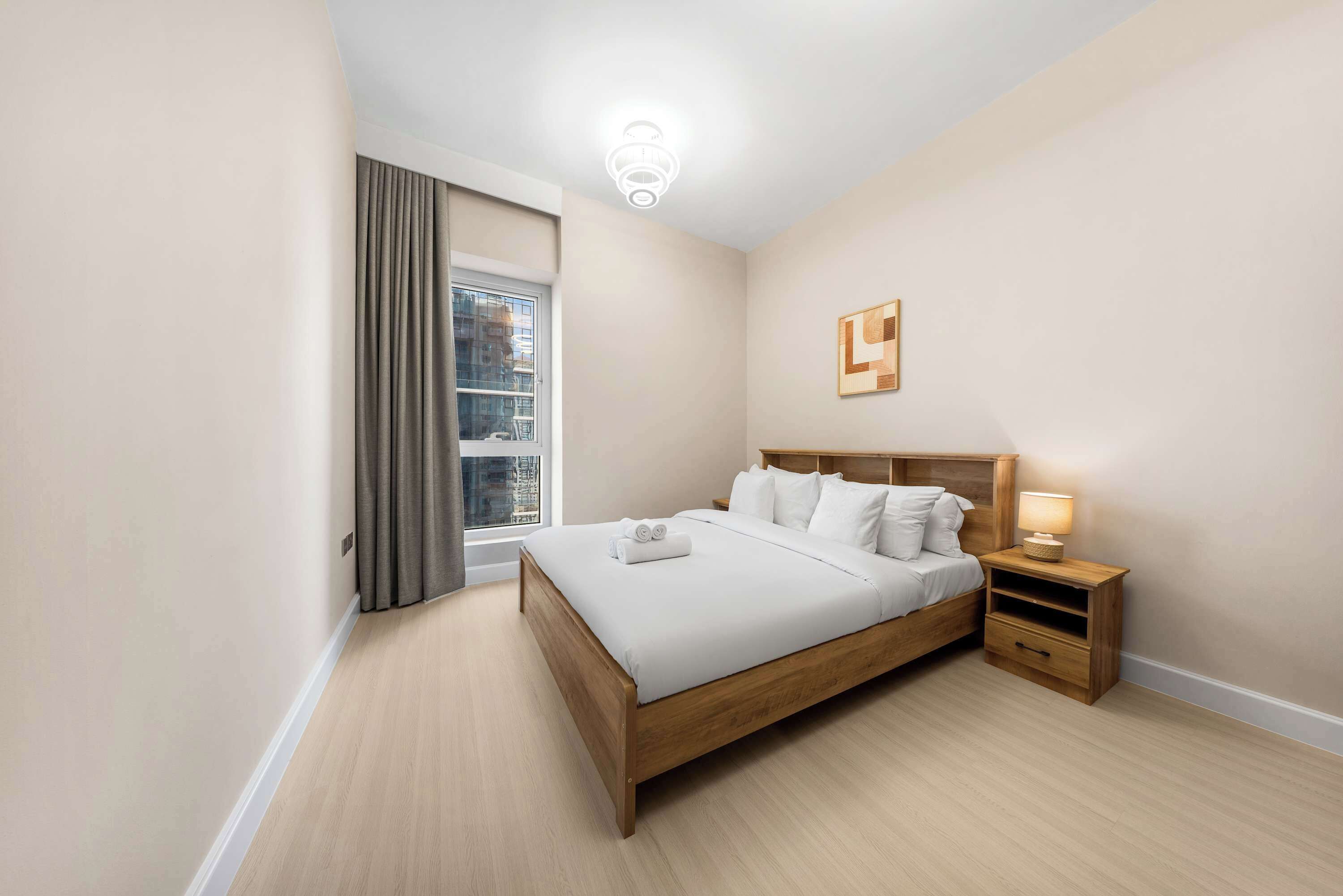A hotel room with a bed, bedside table, lamp, and a view of the city through the window.