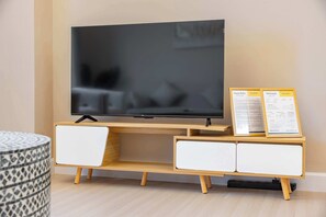 A flat-screen TV on a wooden TV stand with two white cabinets, a small table, and a framed welcome sign. - StayShort - Cozy 2BR Apartment in JLT that Sleeps 4! (JLT)