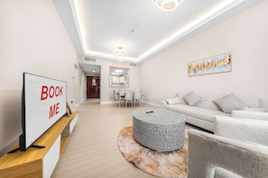 A modern living room with a flat-screen TV on a wooden stand, a sofa with patterned cushions, a round ottoman, and a painting on the wall. - StayShort - Cozy 2BR Apartment in JLT that Sleeps 4! (JLT)