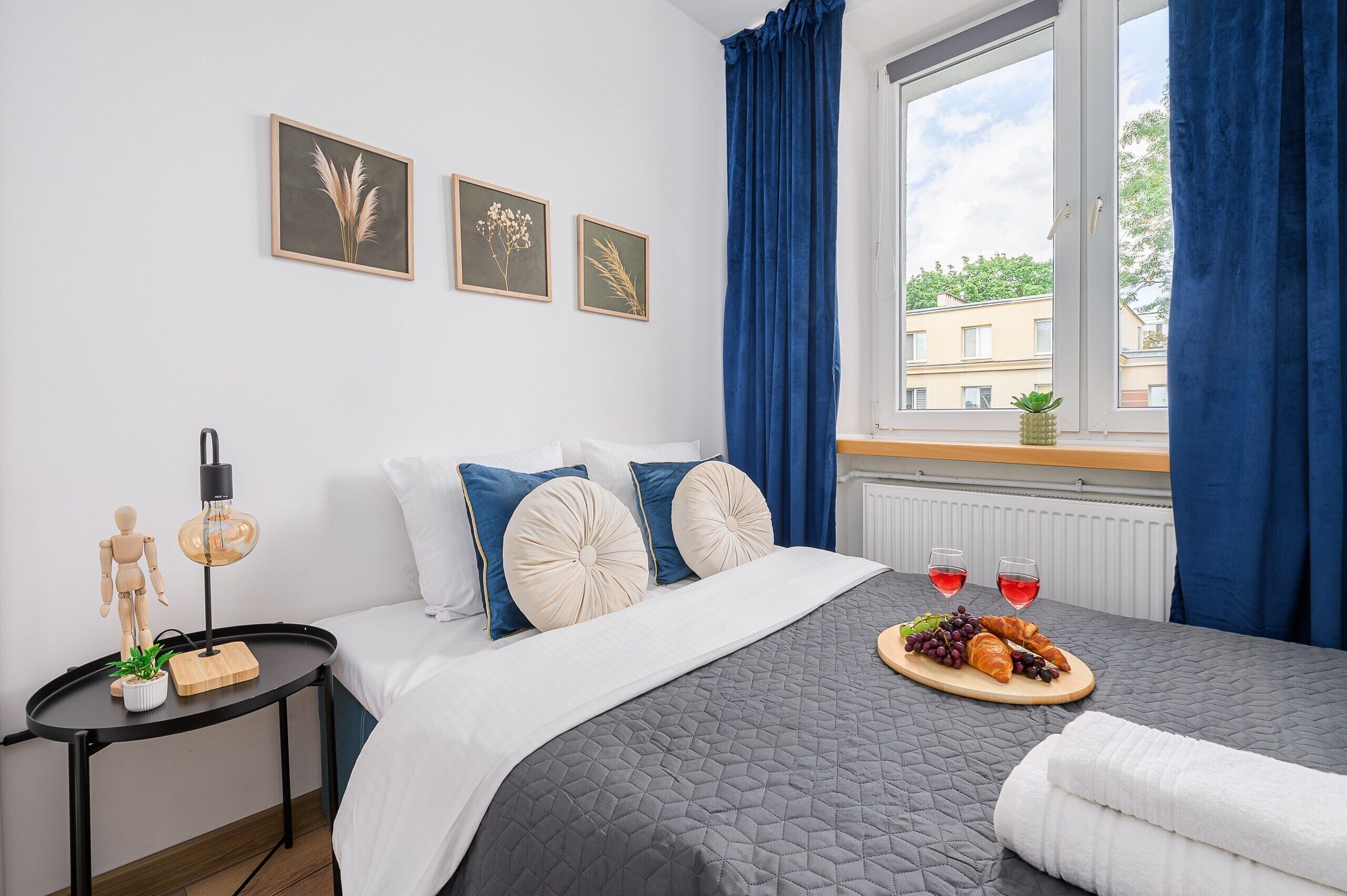 A bedroom with a bed, a bedside table with a lamp, a window with curtains, and a view of buildings outside.