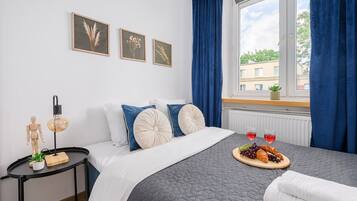 A bedroom with a bed, a bedside table with a lamp, a window with curtains, and a view of buildings outside.