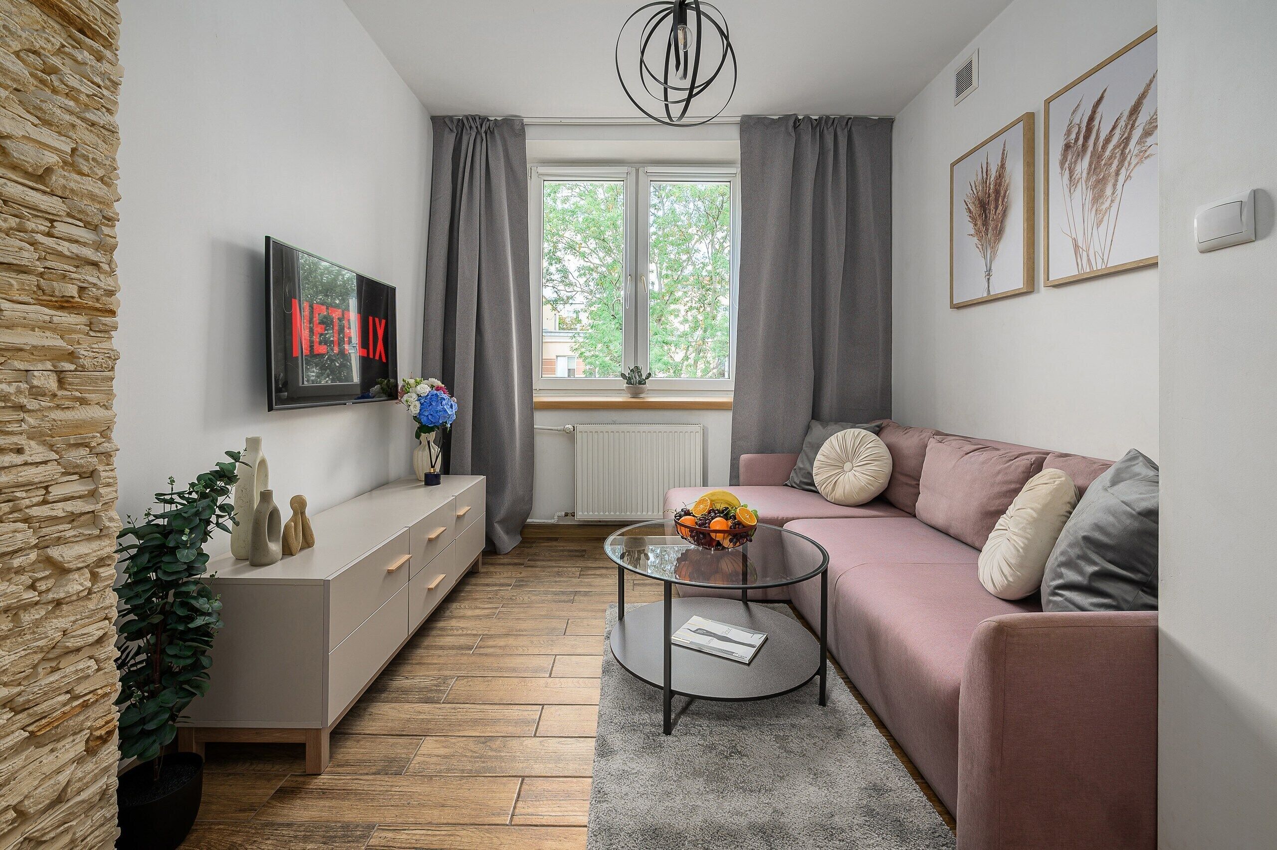 Un salón moderno con una pared de acento de piedra, un televisor de pantalla plana, un sofá rosa, una mesa de centro de vidrio con fruta y una ventana con cortinas.