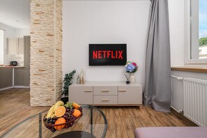 Un salon moderne avec un mur d’accent en pierre, un téléviseur à écran plat affichant le logo « NETFLIX », une table basse en verre sur laquelle sont disposés des fruits, et un meuble aux lignes épurées.