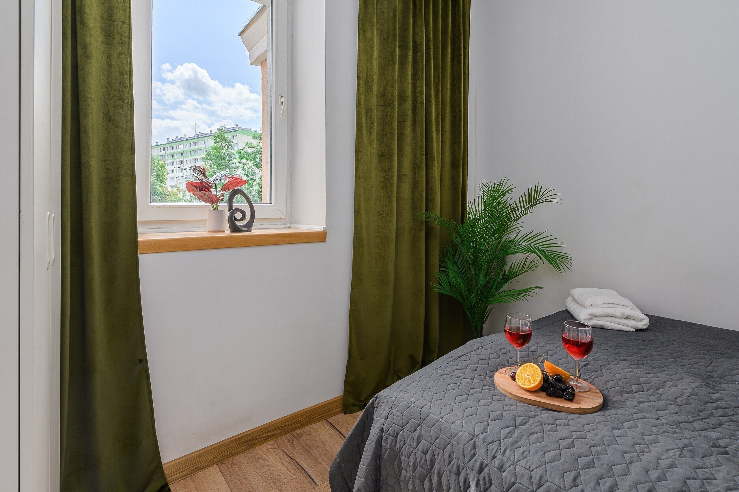 A hotel room with a bed, a window with curtains, a small table with fruit and wine glasses, and a potted plant.