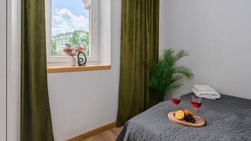 A hotel room with a bed, a window with curtains, a small table with fruit and wine glasses, and a potted plant.