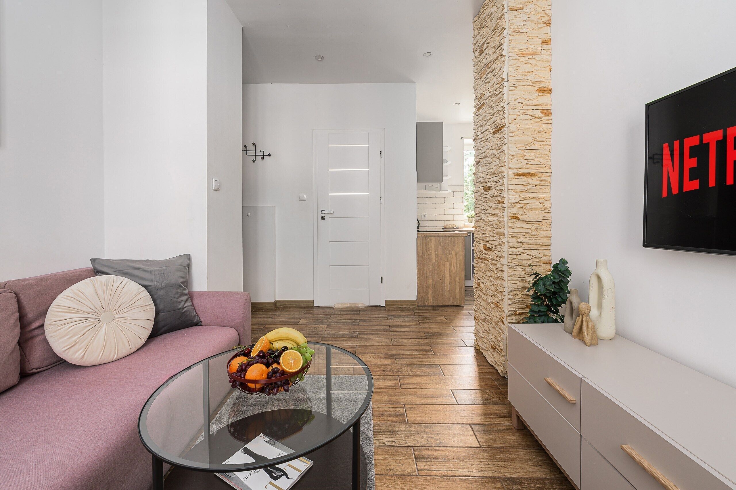 Una sala moderna con un sofá, una mesa de centro de vidrio con fruta, una pared de acento de piedra y un televisor mostrando 'NETFLIX'.
