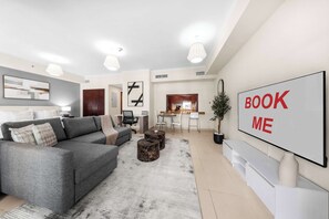 A modern living room with a grey sofa, a coffee table, and a TV screen displaying 'BOOK ME'. - StayShort - Modern and Chic Studio in Downtown with Burj Khalifa Views! (Downtown)