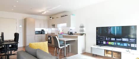 A modern living room with a grey sofa, a dining area with a table and chairs, a kitchen with white cabinets, and a flat-screen TV mounted on the wall.