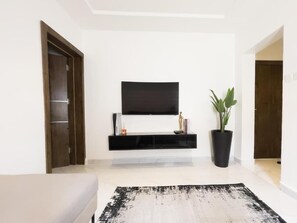 Una sala de estar moderna con un televisor de pantalla plana montado sobre un mueble negro, una planta grande en maceta y una alfombra con textura.