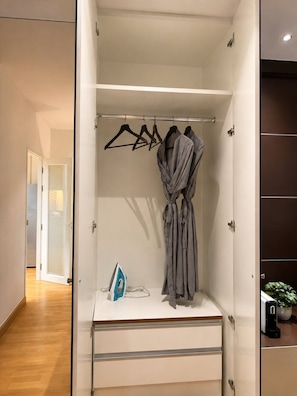 A walk-in closet with a hanging rod, a robe, an ironing board, and a small plant. - One quiet bedroom apartment with amazing balcony views next to Nana and Soi 11 (Khet Khlong Toei)