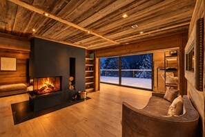 Ein gemütliches Zimmer mit Kamin, Holzwänden, einem großen Fenster mit Blick auf Schnee und einer Sitzecke.