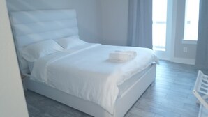 Ein weißes Bett mit Kissen und einem gefalteten Handtuch, ein weißer Stuhl und ein Fenster mit Vorhängen.