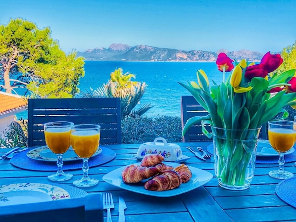 Een gedekte tafel met croissants, een theepot en glazen sinaasappelsap, met uitzicht op een waterpartij en bergen in de verte.